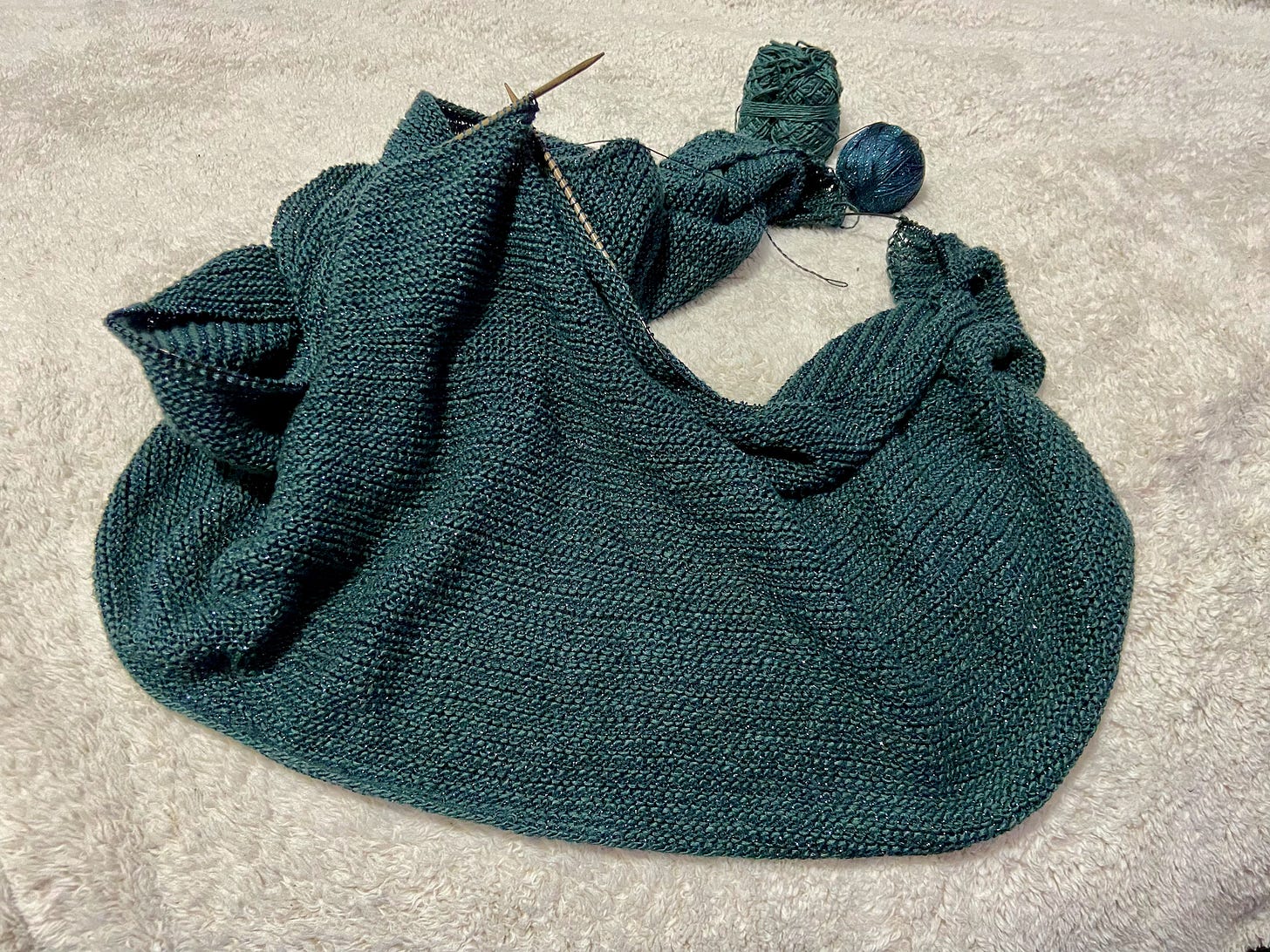 An in-progress Mineral Wrap, made with two different shades of blue yarn, attached to knitting needles. It's propped up and placed on an off-white throw blanket.