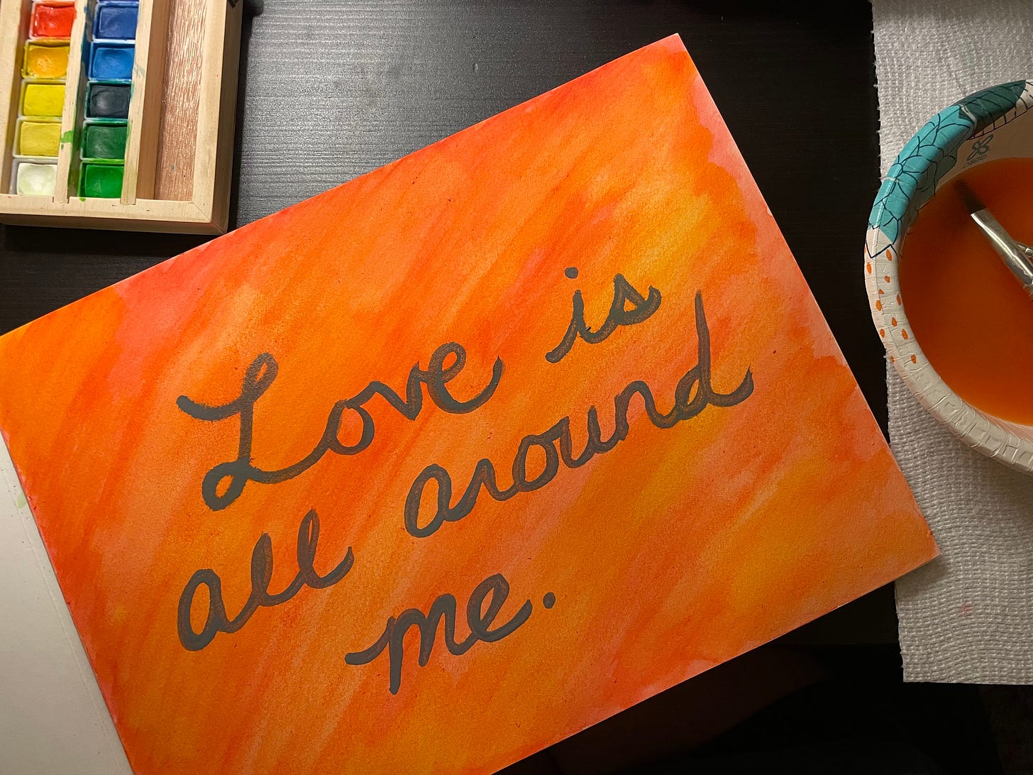 A watercolor painting with a red, orange, and yellow background. "Love is all around me." is painted in gray letters.