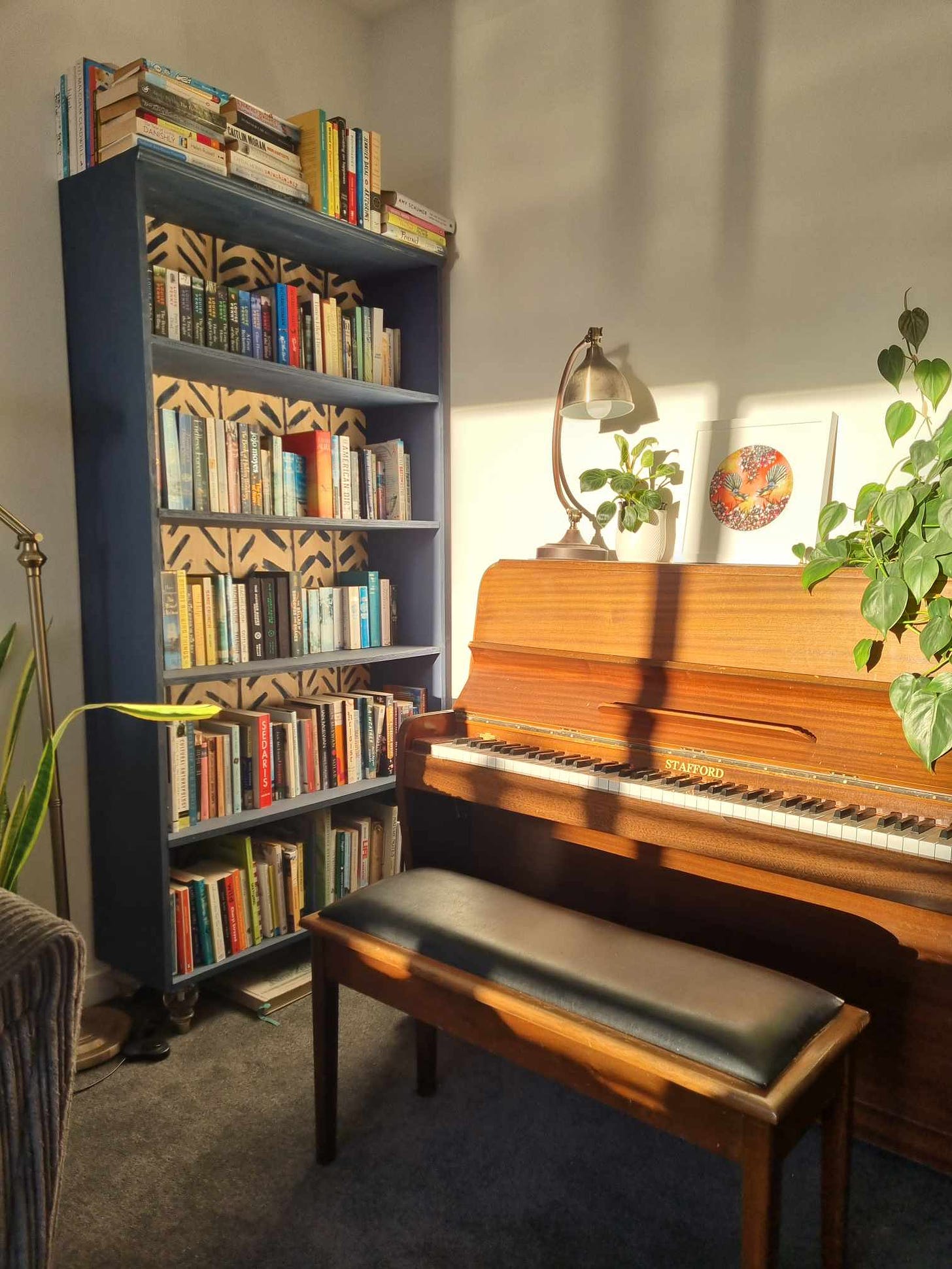 A corner of Charlotte's house with a piano, large bookshelf, and some houseplants, glowing golden in the morning sunshine.
