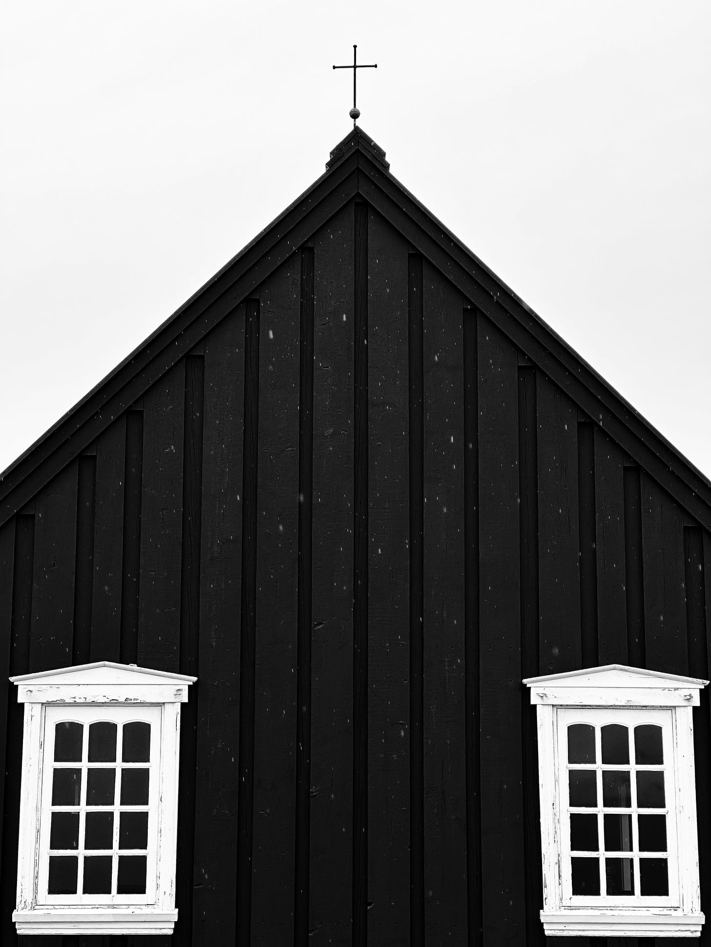 Búðakirkja - The Black Church of Budir a close up photograph of the back of the church.  The weathered, white painted windows are shown on the bottom left and right corners.  The black siding stretches up to the point of the roof and the simple metal cross is silhouetted against the light gray sky.