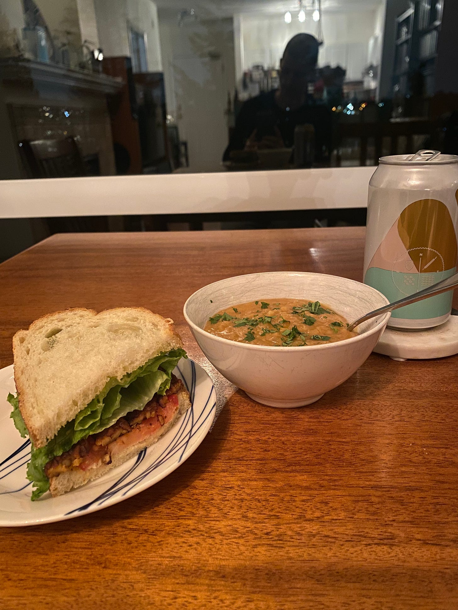 A side plate with half a tempeh BLT on it, next to a little white bowl of soup sprinkled with cilantro. A can of Four Winds lager is on a coaster on the right side of the bowl. The window is dark in the background.