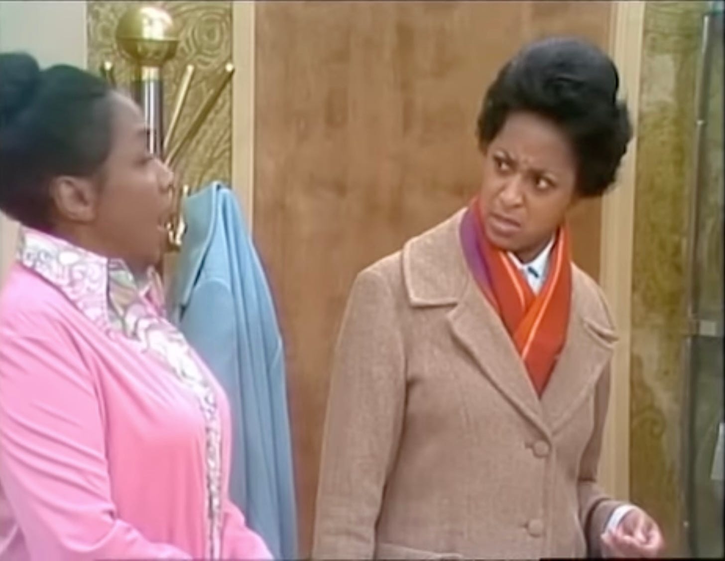 Louise (Isabel Sanford) and Florence (Marla Gibbs) interact in the pilot episode of the Jeffersons. Two Black women, one in a pink sweater and the other in a tan coat look at each other