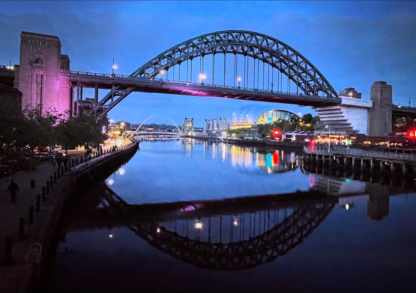 A view from Newcastle/Gateshead's Swing Bridge, up the Tyne to the Tyne Bridge and the Millennium Bridge just visible in the distance. The water is still and the bridges are reflected, the sky is a blue evening light, and the riverside buildings are lit in the dusk.