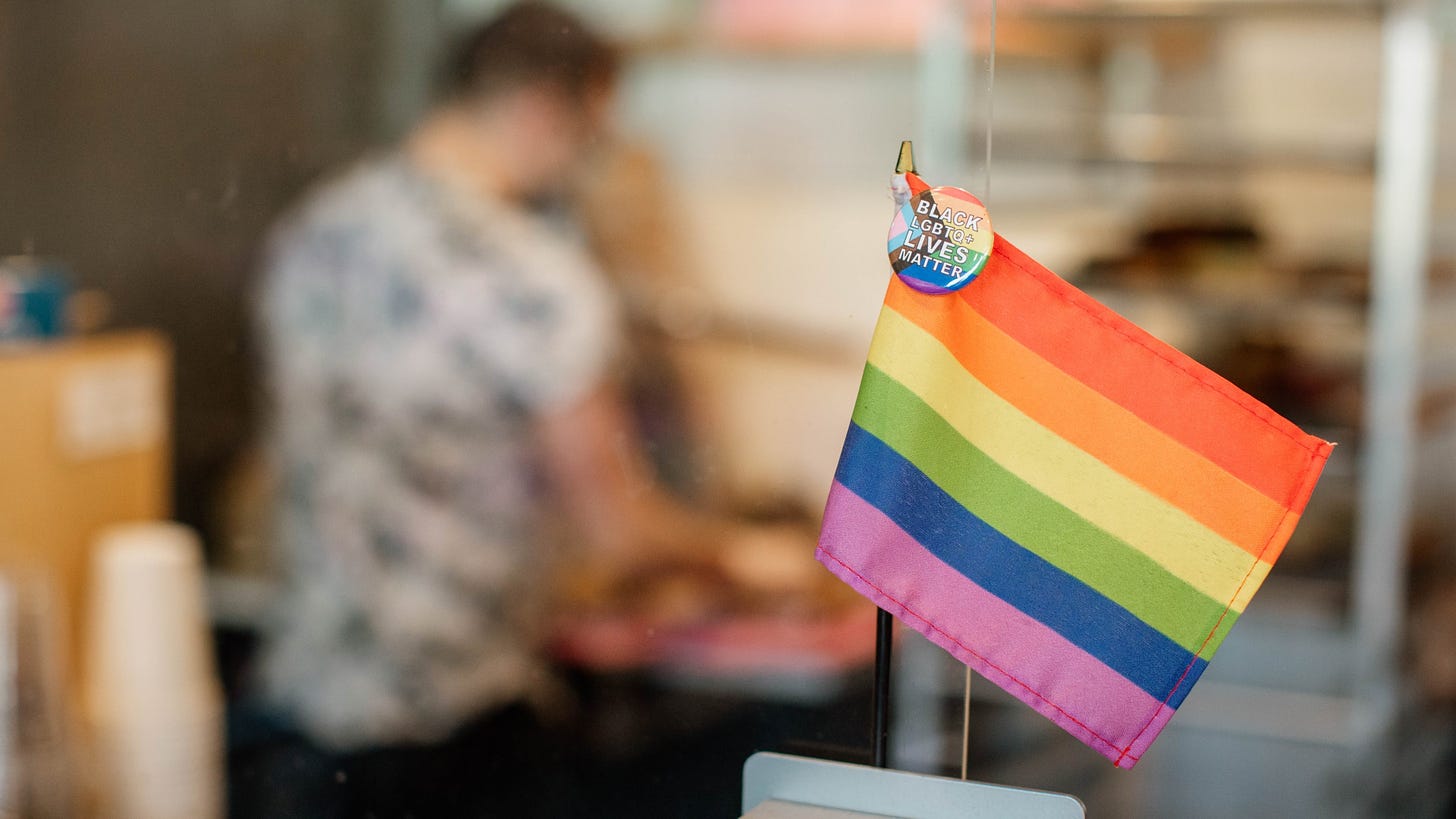 A Pride flag on the register at Strange Matter Coffee, with an out of focus employee and takeaway cups in the background