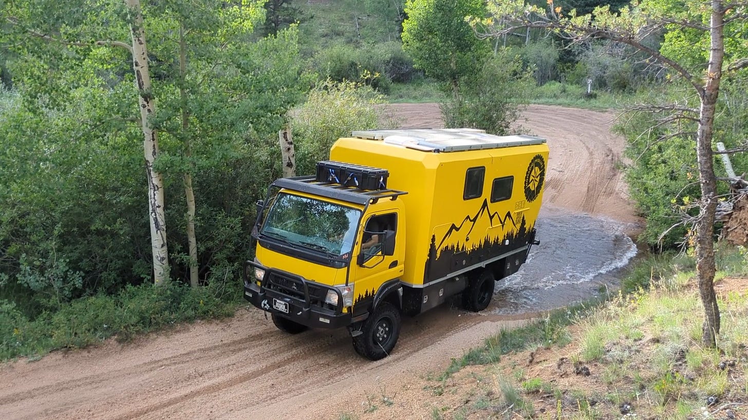 Walter the big yellow truck, in a beautiful wooded setting, having just crossed a stream, driving on a red dirt road