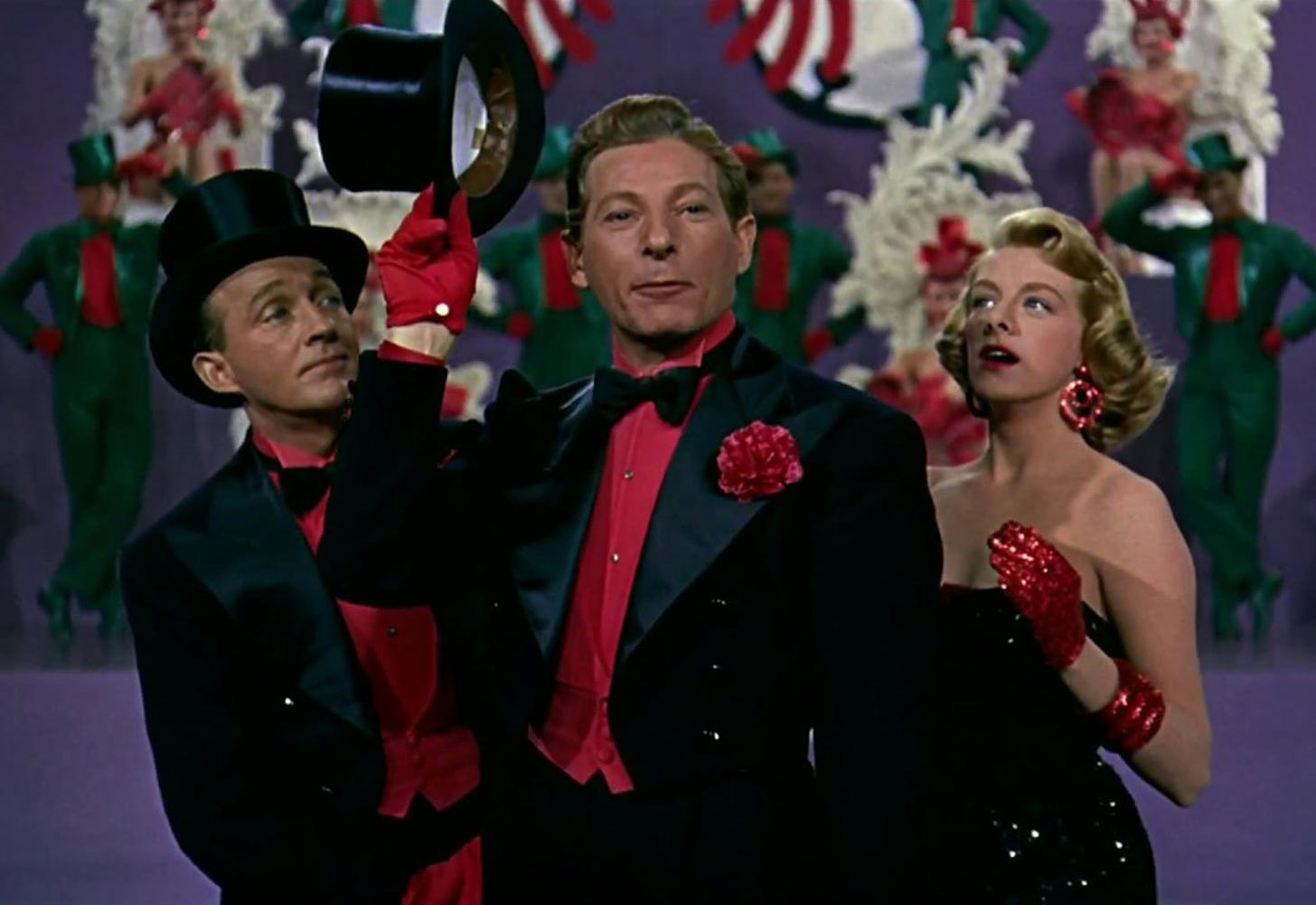 A still from a dance number in the movie White Christmas, featuring Danny Kaye, Bing Crosby, and Rosemary Clooney. Kaye and Crosby are wearing tuxedos with red shirts, red gloves and black top hats. Clooney is wearing a black dress with red gloves. 