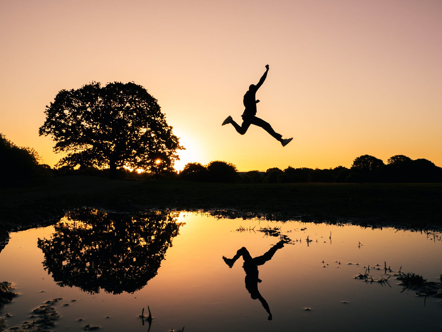 The silhouette of a person jumping over a pond. The sun rises in the background. The sky is a peach color, with the water reflecting the same color.