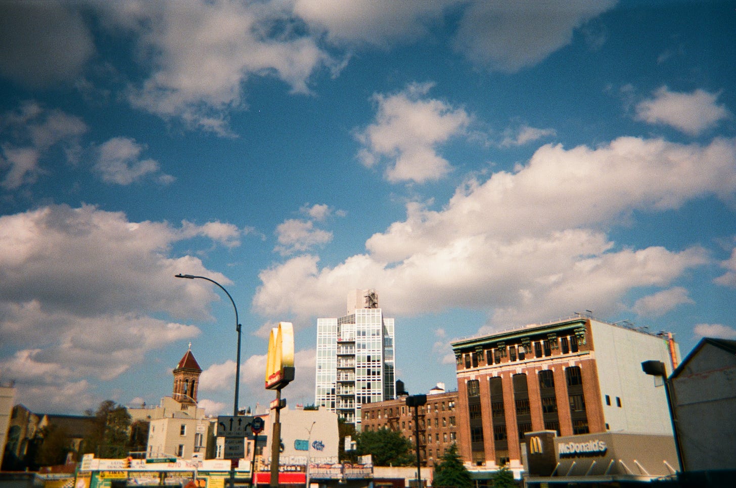 A skyline with a McDonald's, an old bank building, and a new tower crowned by a massive, open sky with fluffy clouds.