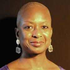 Photo of Eva Yaa Asantewaa, a Black woman with bald head and crystal cube earrings smiling at the camera against a dark background.