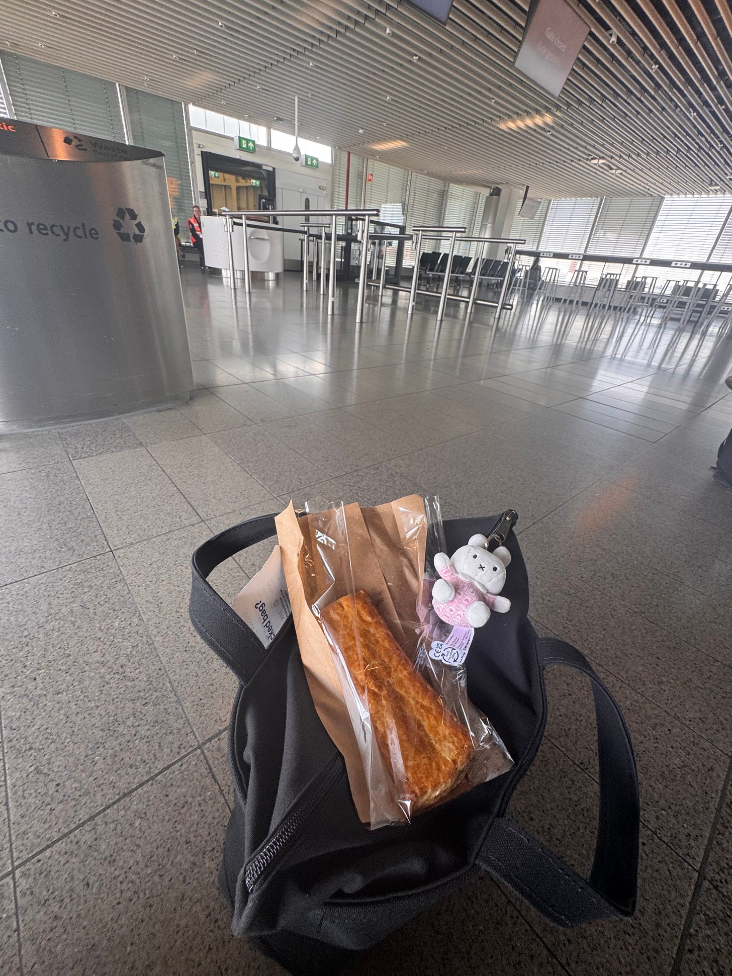 Black bag on airport floor with a packaged pastry and pink bunny keychain, set against a quiet terminal with empty seats and a recycling bin nearby.