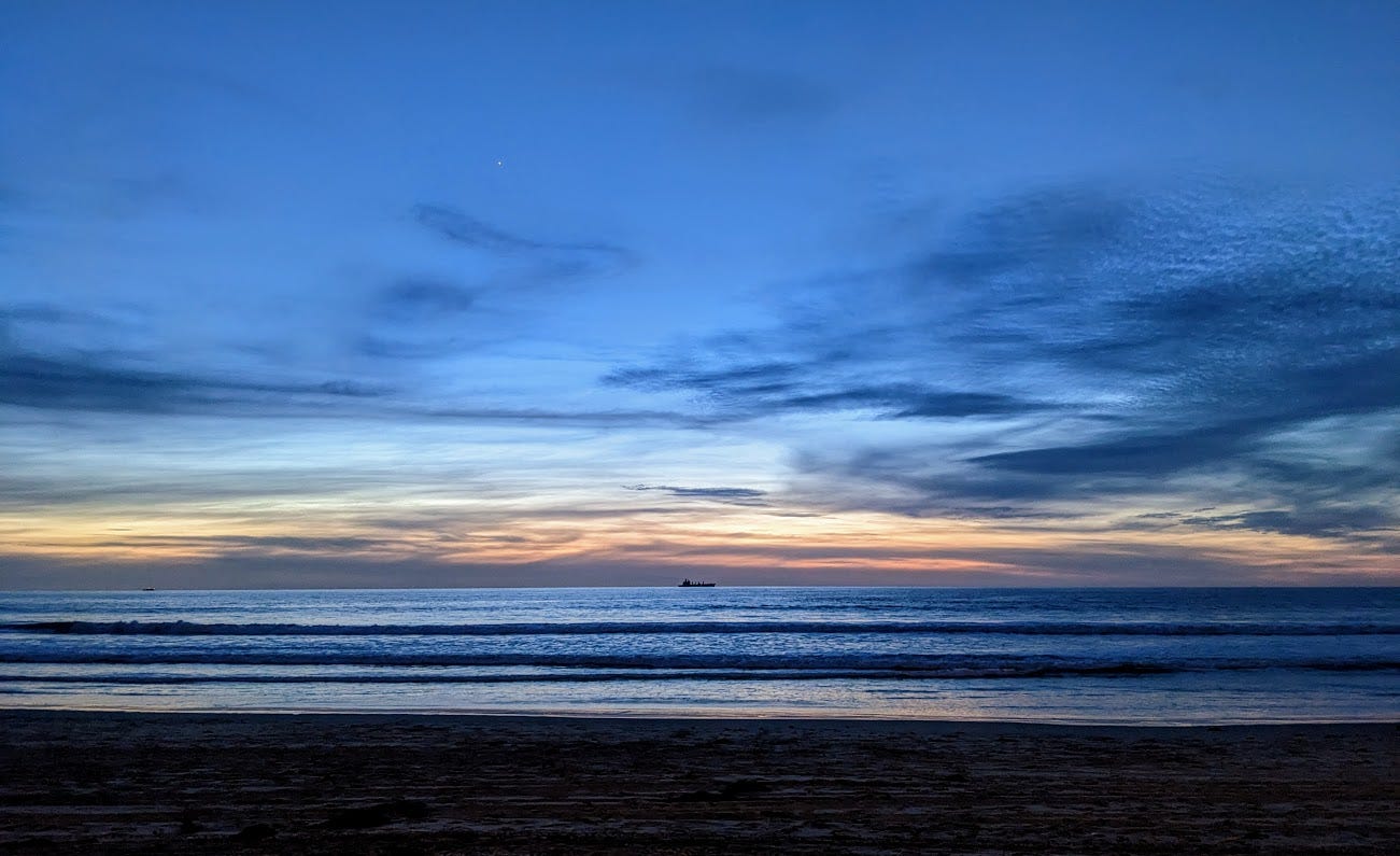 A freighter sits on the Pacific Ocean of Mission Beach in the center of the picture. The freighter is sitting on top of the blue ocean, with rolling waves heading toward the dark sand in the foreground. Along the horizon, level with the freighter, there are pink and purple clouds below an electric blue sky
