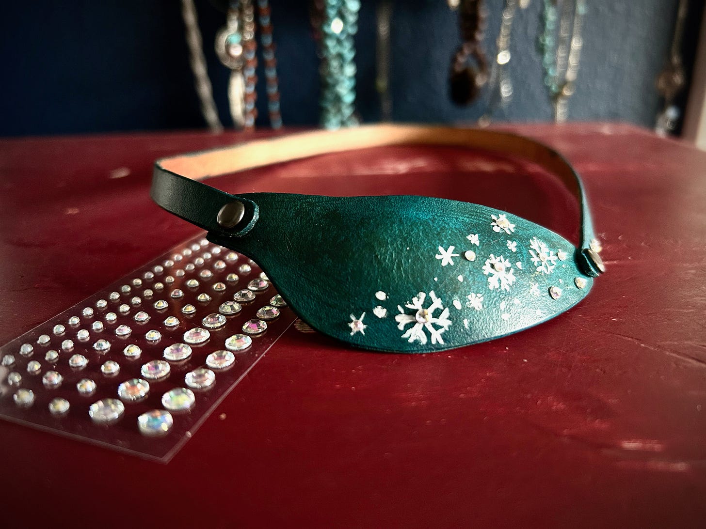 Closeup of a blue eye patch sitting on a dresser. White snowflakes are painted on the eye patch, with rhinestones in the middle of the flakes
