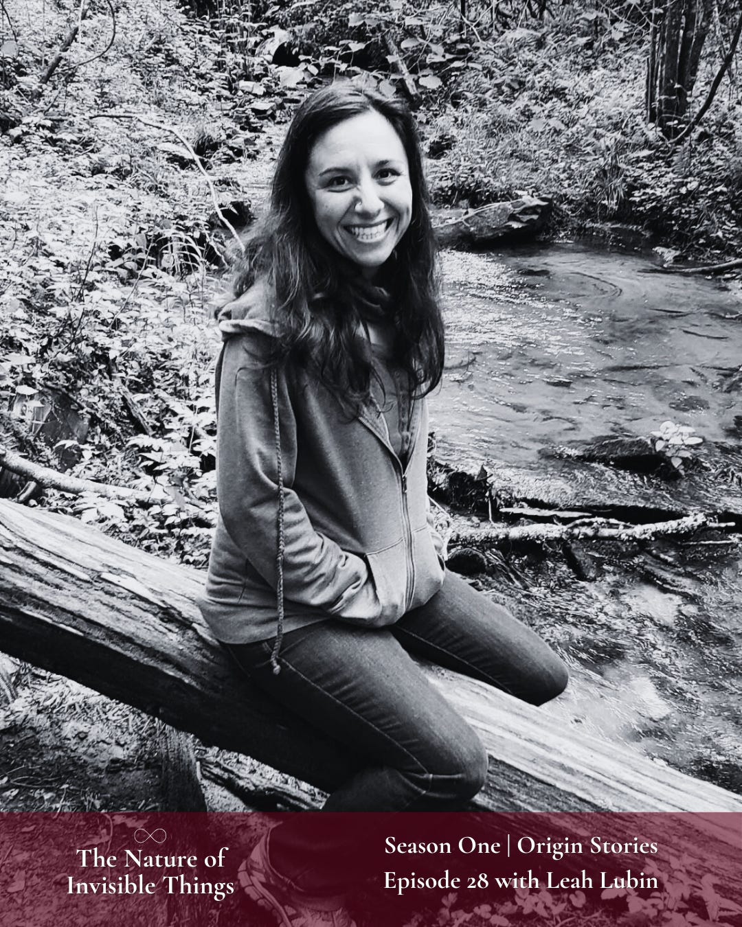 Leah, a woman with long dark hair, is sitting astride on a fallen log near a stream in the woods. She is wearing a zip-up hoodie and denim jeans while smiling brightly at the camera.