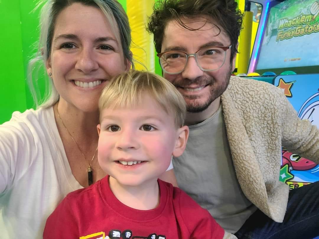 A white family of three posing for a group selfie in an arcade. The mom and dad are middle-aged with big smiles, having fun but also a bit run down from the week. In front huddled between them is their 4 year old son, pink cheeks flush from running around and playing arcade games with his cousins. He has a big smile and is starry eyed from all the flashing lights and sounds from the games. 