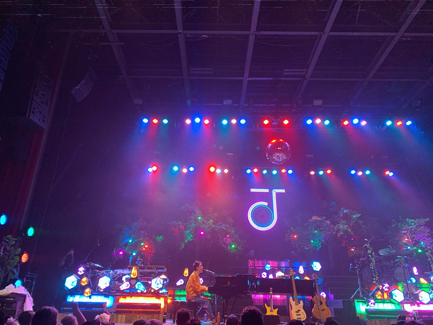 Jacob Collier, a musician from the UK, playing the piano and singing at the Coca-Cola Roxy venue. Jacob is wearing a flashy orange shirt, funky pants, and there are bright, colorful lights everywhere.