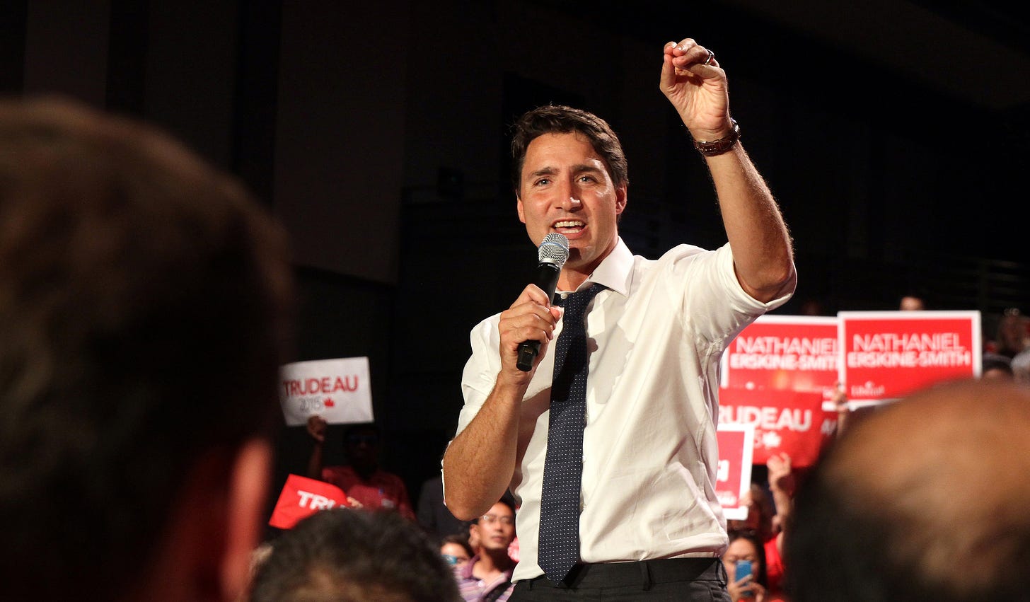 Justin Trudeau raises his hand and holds a mic at a campaign rally with Liberal signs in the background.