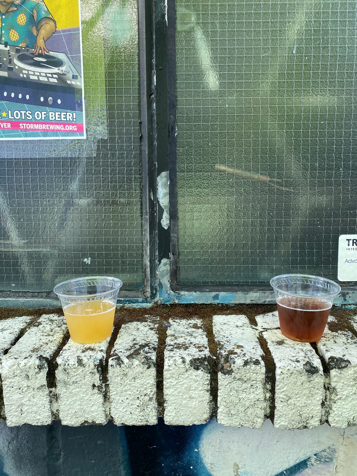 A brick window ledge outside Storm Brewing. The bricks have moss growing in between, and on the window a corner of a poster advertising a party with 'lots of beer!' is visible in the upper left. Two small plastic cups of beer sit on the ledge, one cloudy IPA and one darker ale.