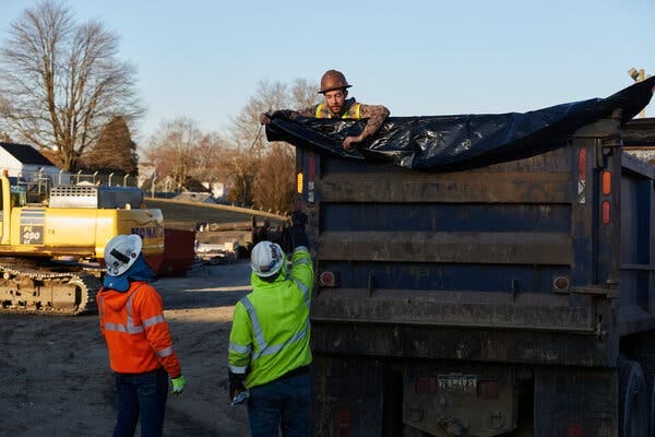 Workers in fluorescent yellow and orange gear stand in front of a truck bed. One of them gestures to another worker in a hard hat, who is on the truck.