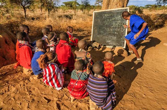 Children sit in front of a chalkboard under a tree outdoors