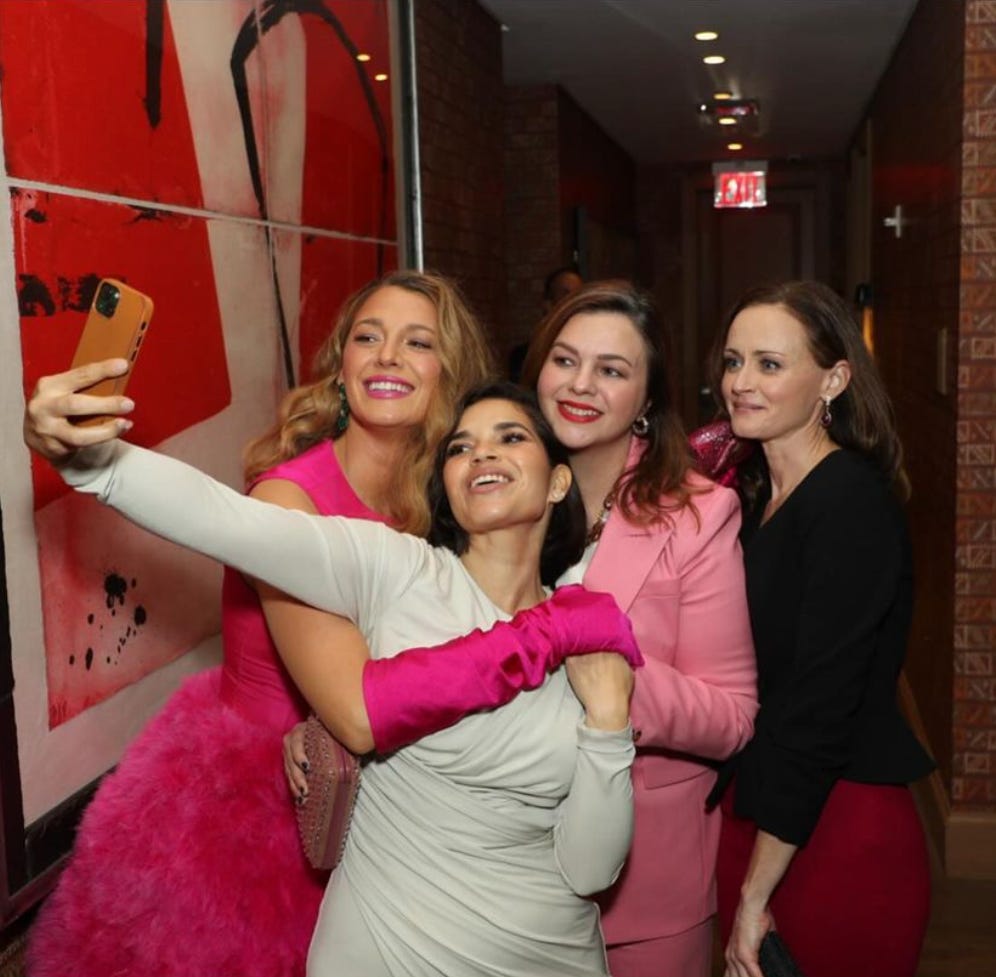 A photo of Blake Lively, America Ferrera, Amber Tamblyn, and Alexis Bledel taking a group selfie. America holds her phone out to take the photo. Everyone has their arms wrapped around each other as they smile into the camera.