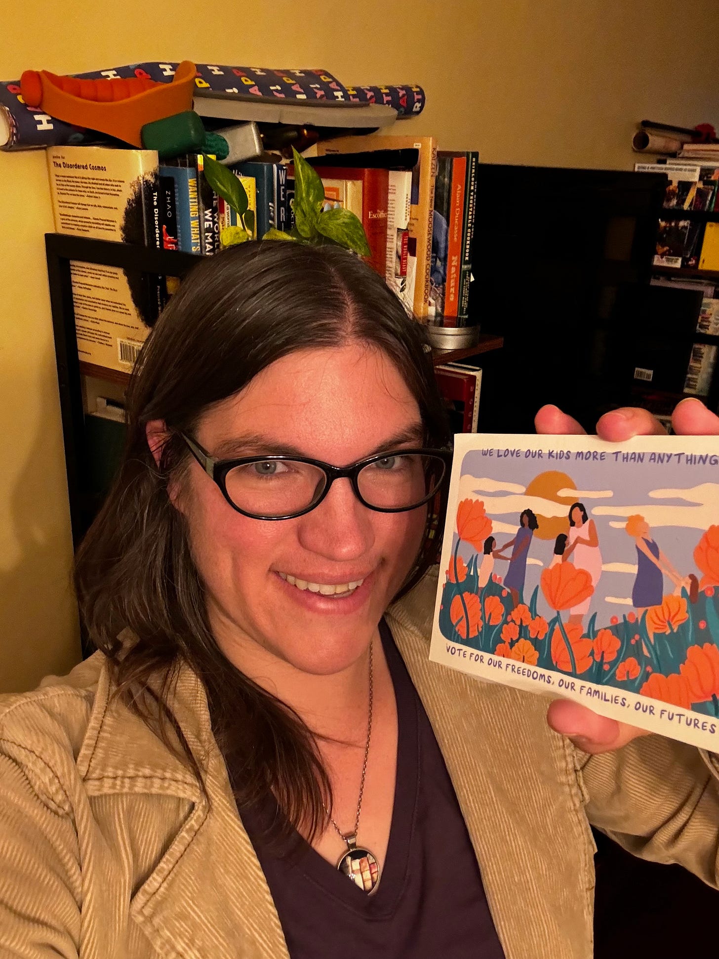 Me (a white woman in a tan jacket) holding up a postcard with an illustration on it to encourage voting, standing in front of a messy bookshelf