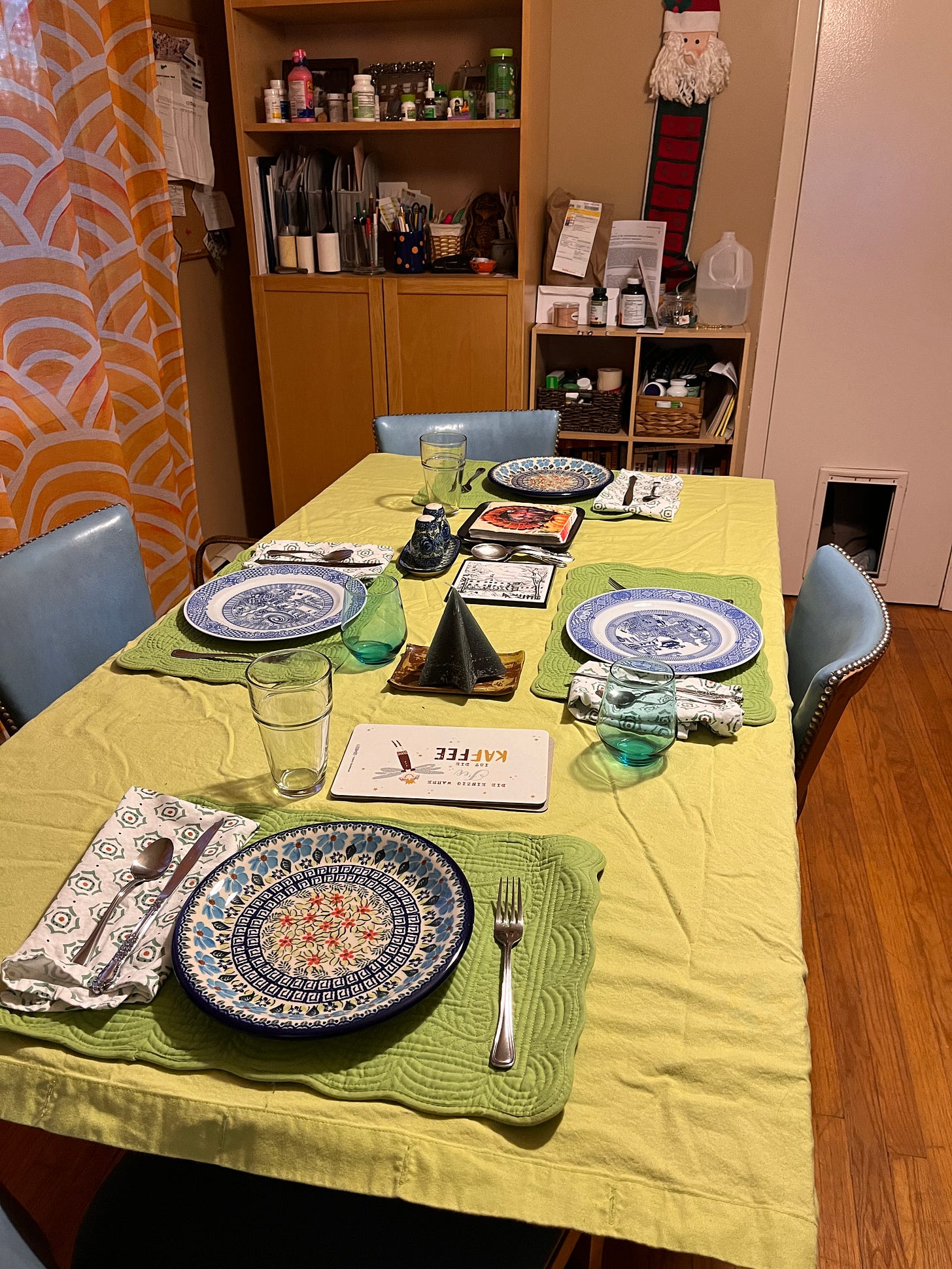 A table set with a mix of blue and white plates on a lime-green cloth, sorta wrinkly, on green placemats, four chairs, with a shelf behind it cluttered with pill bottles and papers, and a curtain hanging to the left that is gauzy orange.