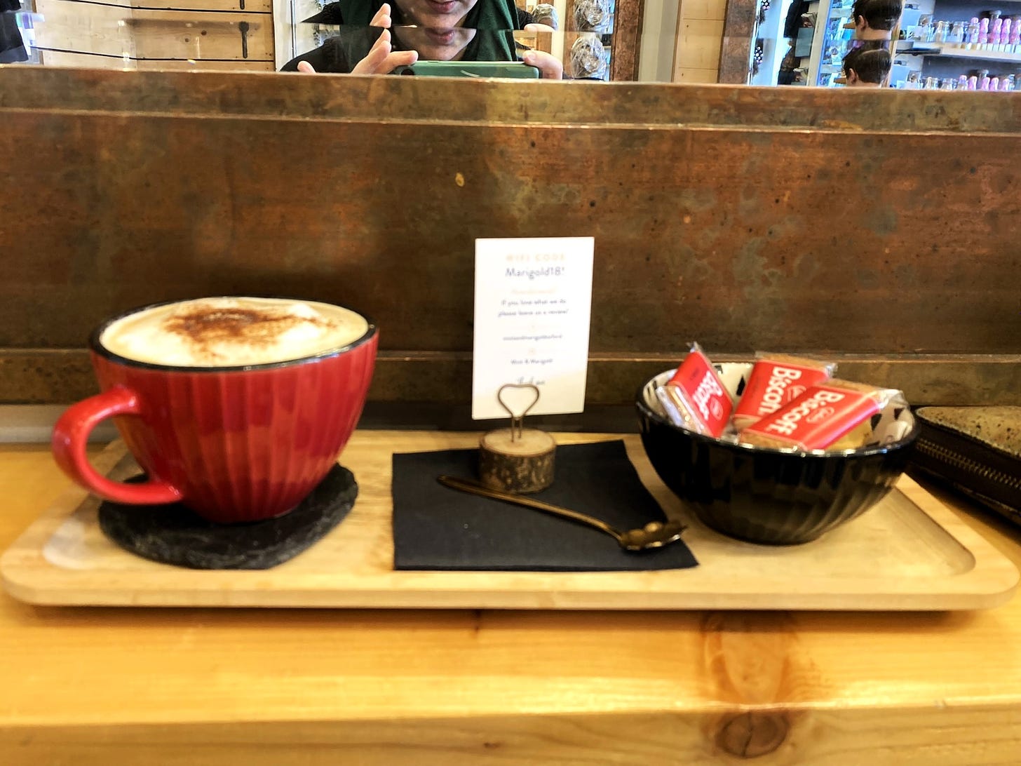 A large red mug contains a very frothy cappuccino. To the right is a black bowl containing biscoff biscuits. 