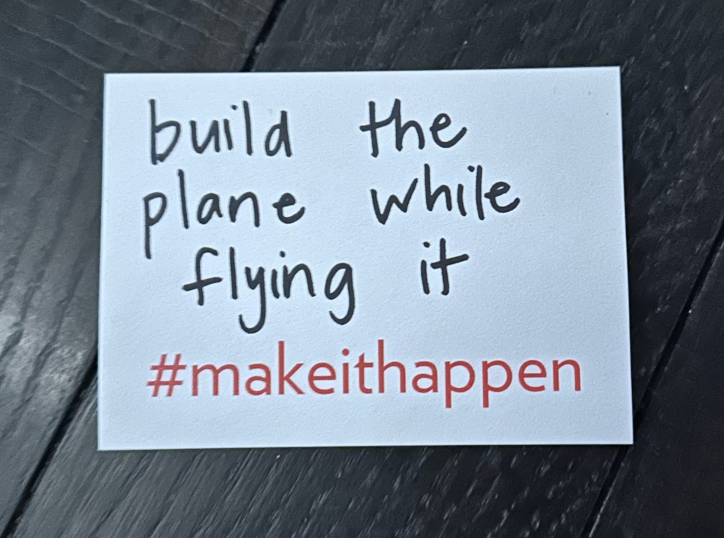 a photo of a sticky note that has, “build the plane while flying it” written on it and includes #makeithappen preprinted on the bottom
