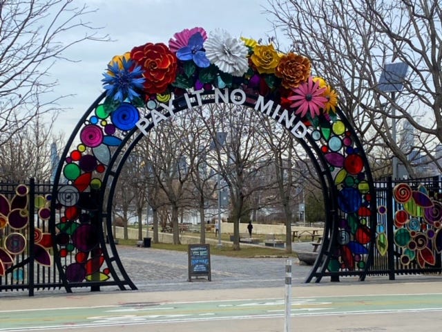 The arched gateway to the Marsha P. Johnson State Park. It has a stained glass effect and is crowned in flowers as she wore. Beneath in white block letters it reads, "Pay It No Mind."