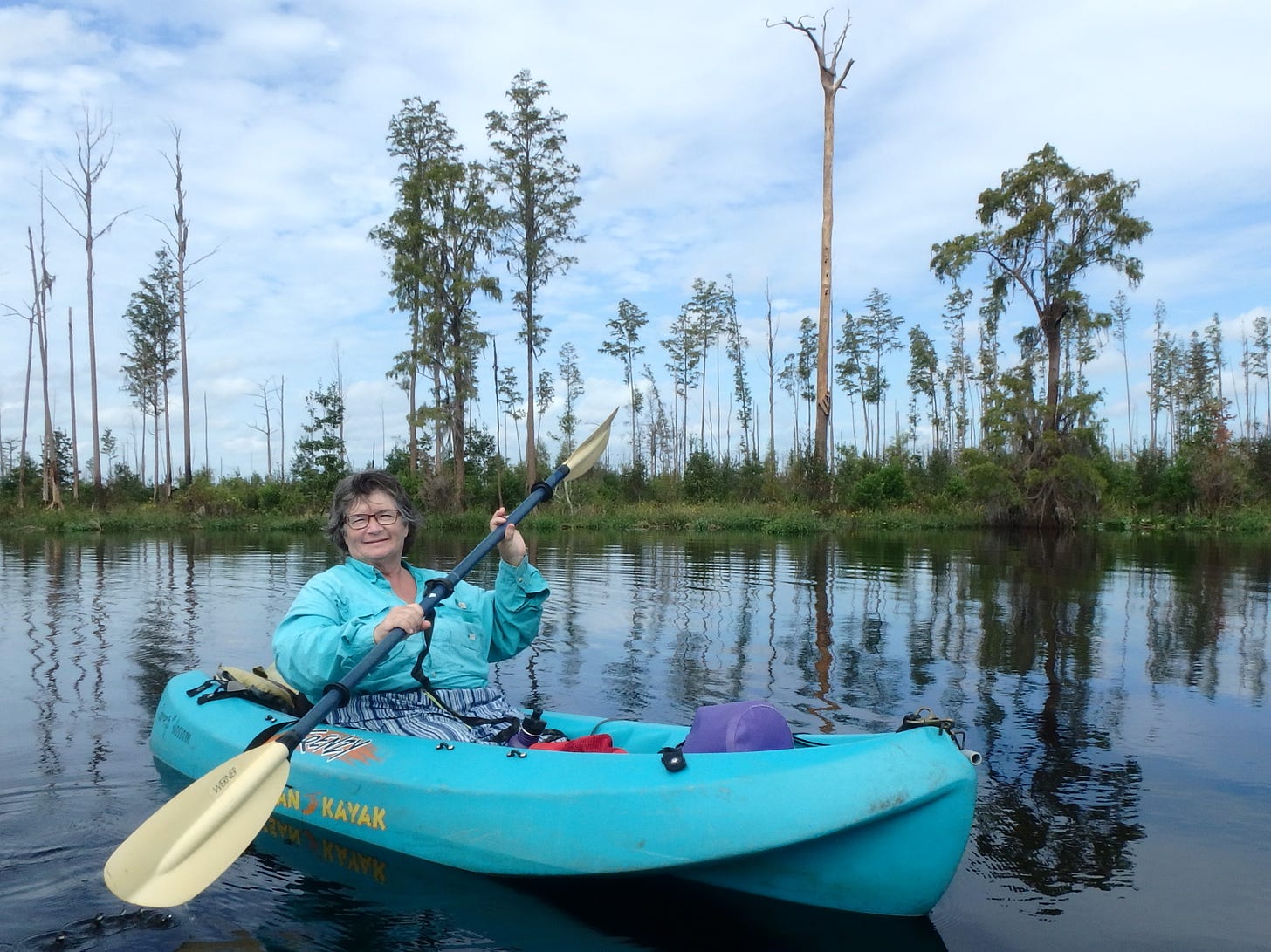 An older white woman (the author) paddling in a blue kayak in an open area of a swamp with low bushes and a few tall cypress trees in the background. 
