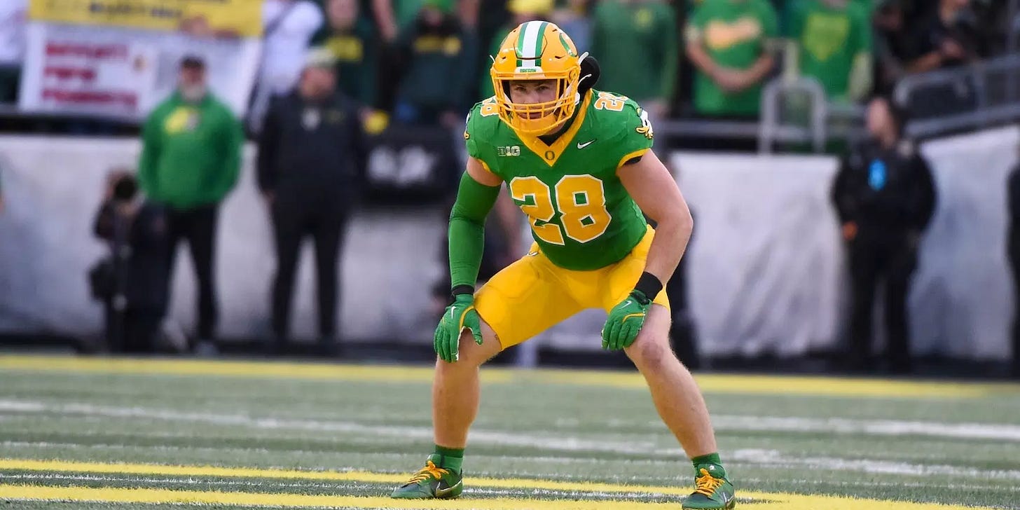 Oregon Ducks linebacker Bryce Boettcher wearing a green jersey with yellow pants, number 28, in a defensive stance on the field during a game. Fans and sideline staff are visible in the background.