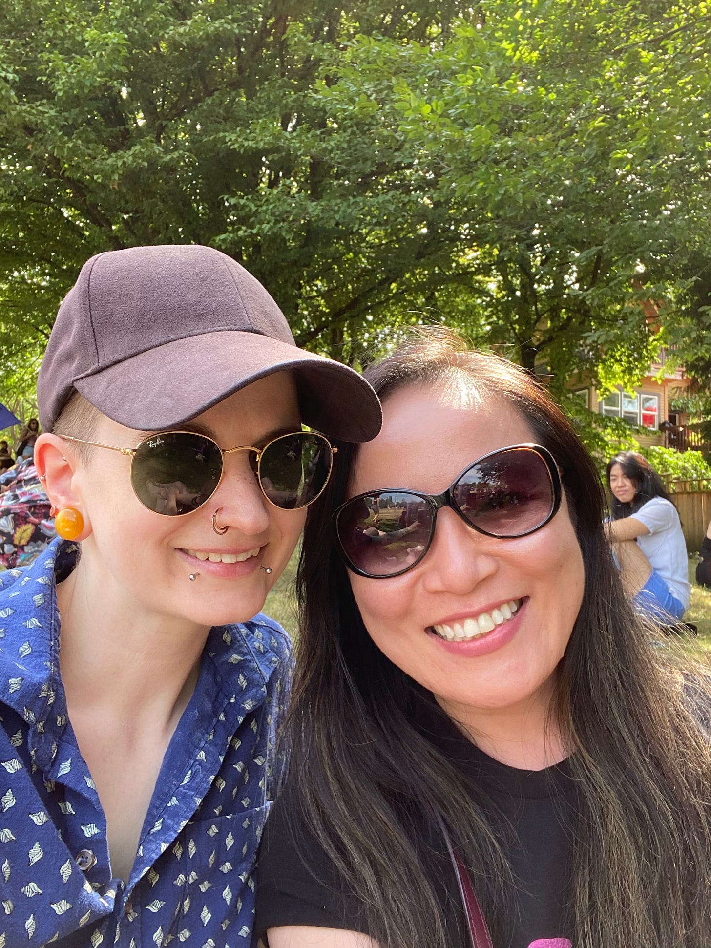 A photo of Liang and me at McSpadden Park before the march. We both look into the camera, smiling. Liang's long dark hair hangs over her shoulders and she is wearing sunglasses and a black t-shirt. I am wearing a black baseball cap, sunglasses, and a blue shirt with a seashell pattern, two of the top buttons undone. The background of the photo is full of trees.