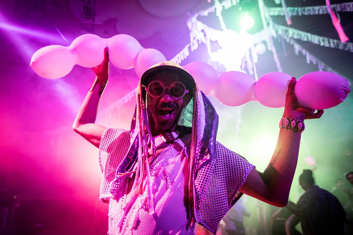 A person smiles directly at the camera as they hold up balloons resembling two ponytails on their head. They are wearing a white mesh top, white circle glasses and a had with fringe accessories on