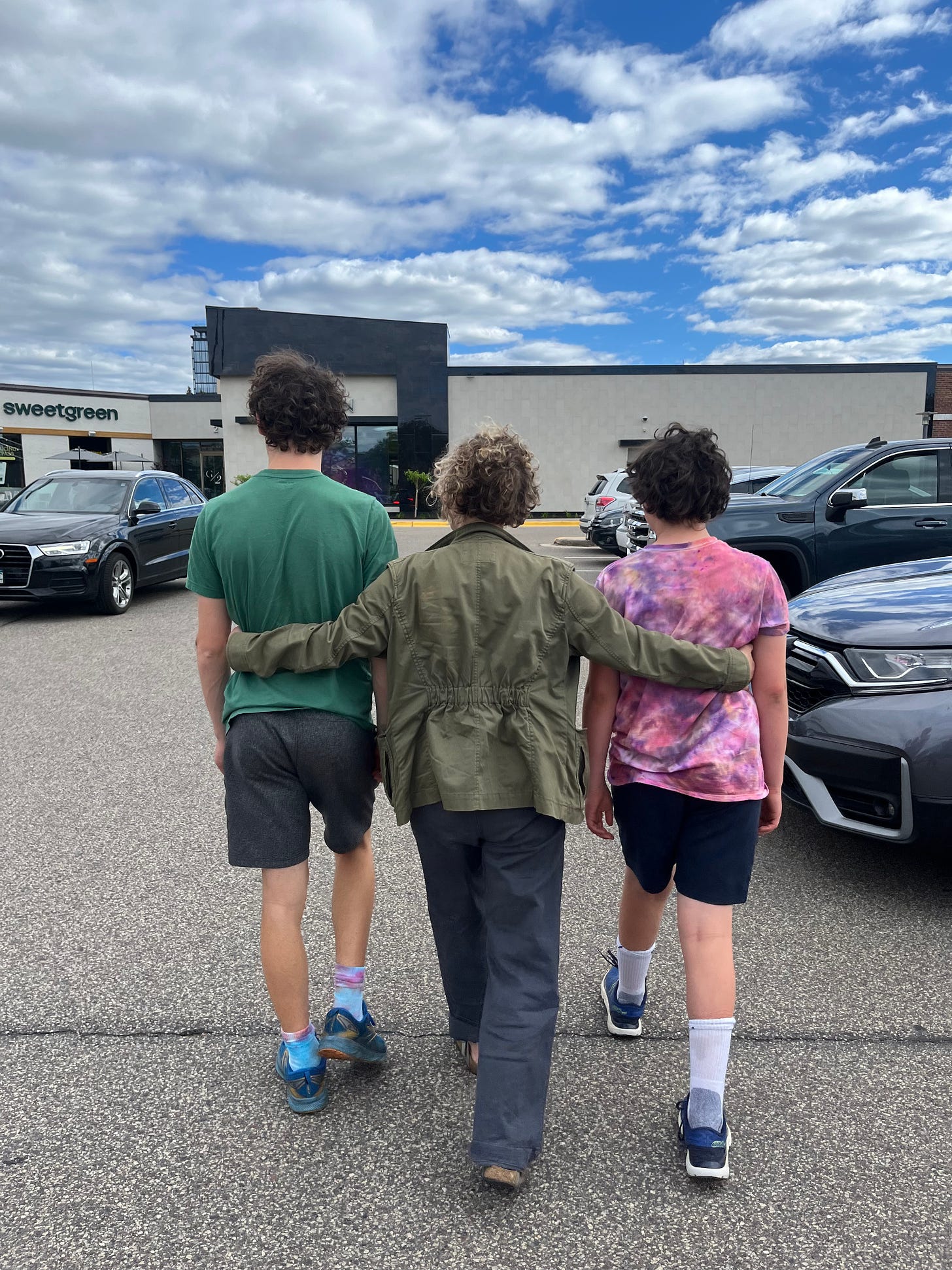 a mother with her arms around her tween and teen son walk through a parking lot