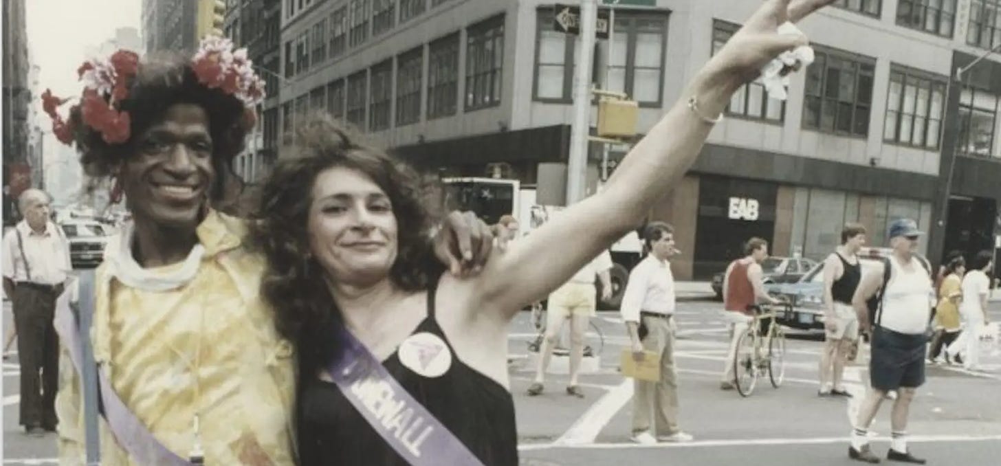 Marsha P. Johnson, wearing a yellow dress and a crown of flowers, holds Silvia Rivera, in a black dress, as she flashes a V sign with her left hand.