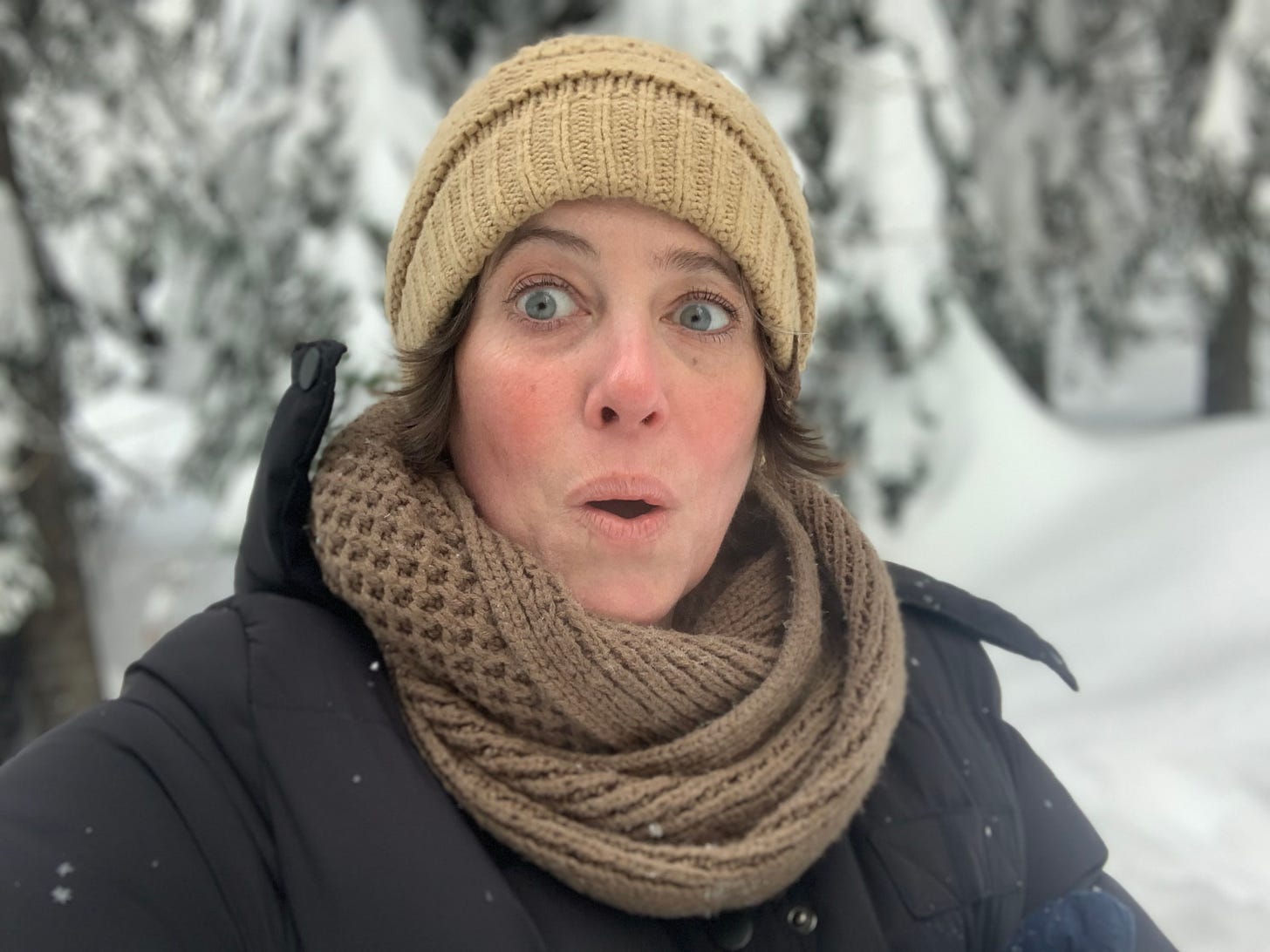 Author Holly Starley, her eyes playfully wide, stands in a snow-covered field of pine trees.