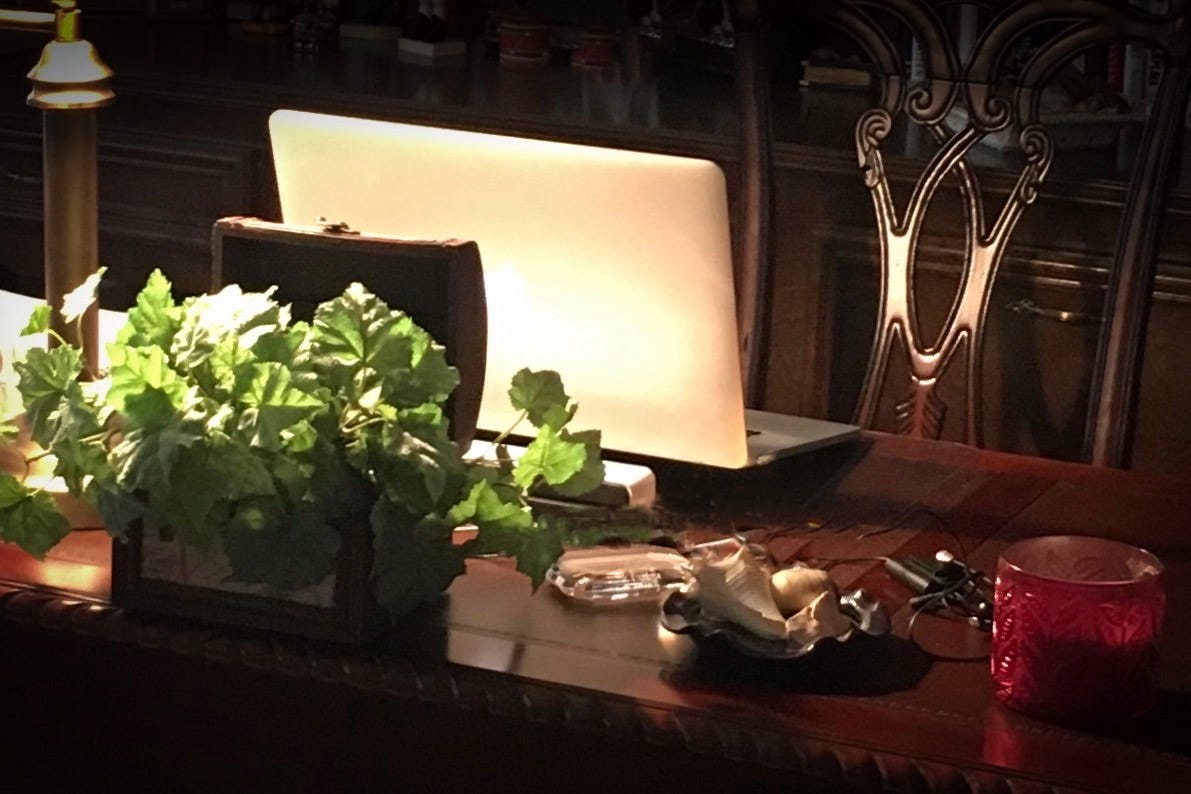 Desk with a plant, computer, a red candle holder, and an antique chair behind it with light from a lamp shining from the left of the photo