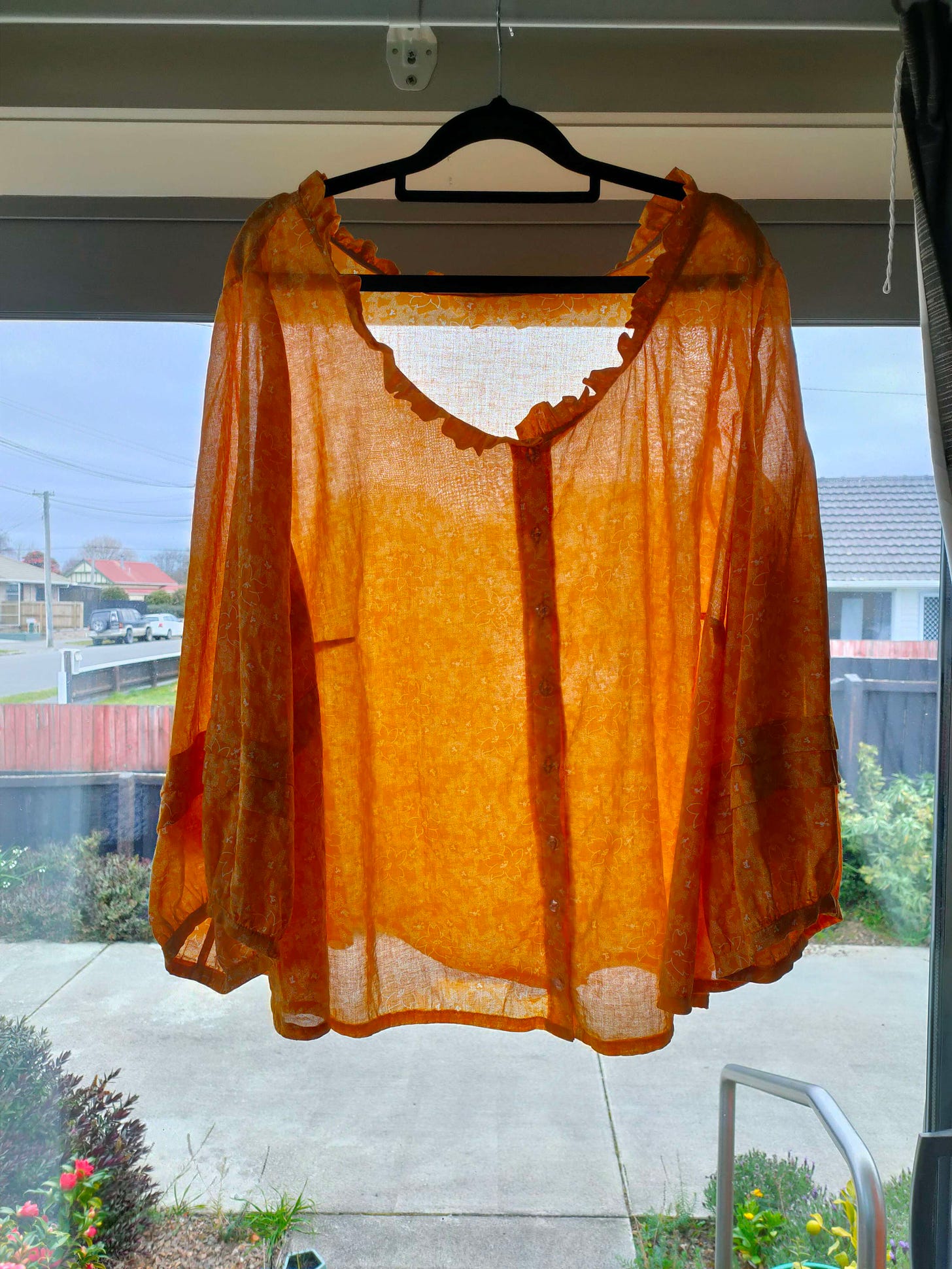 A yellow-orange blouse, semi-translucent against the light, hangs from a clothes hanger hooked over a curtain rail. The blouse has a ruffled neck that took approximately forever, and tuck sleeve details that are barely noticeable because of the lovely floral pattern, but I'm sill very proud of how neatly I managed them.