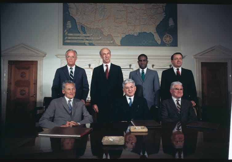 Three men in suits sit side by side at a conference table while four men, also wearing suits, stand behind them. A large map of the US hangs on a wall in the background.