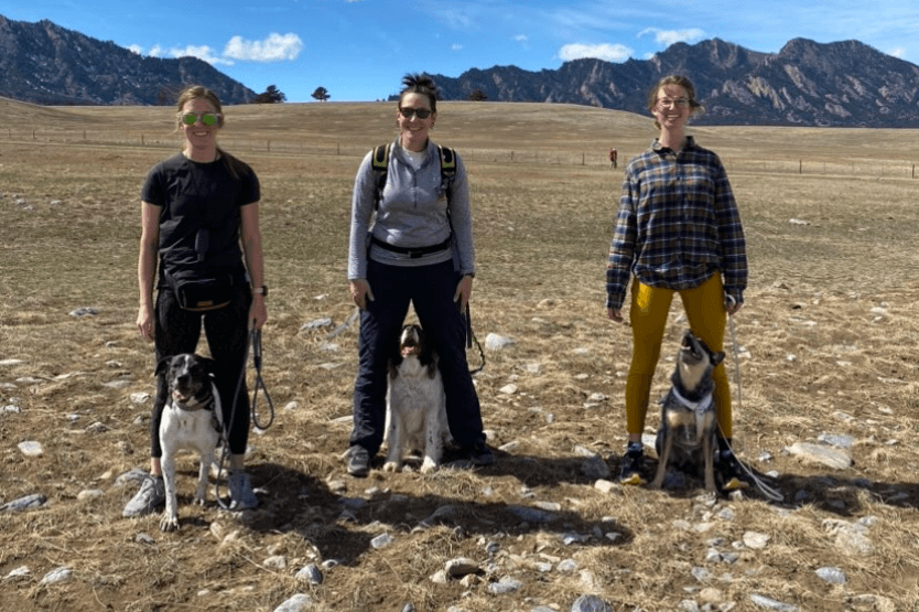 Three women stand on a Colorado trail, each with their dog posing between their legs