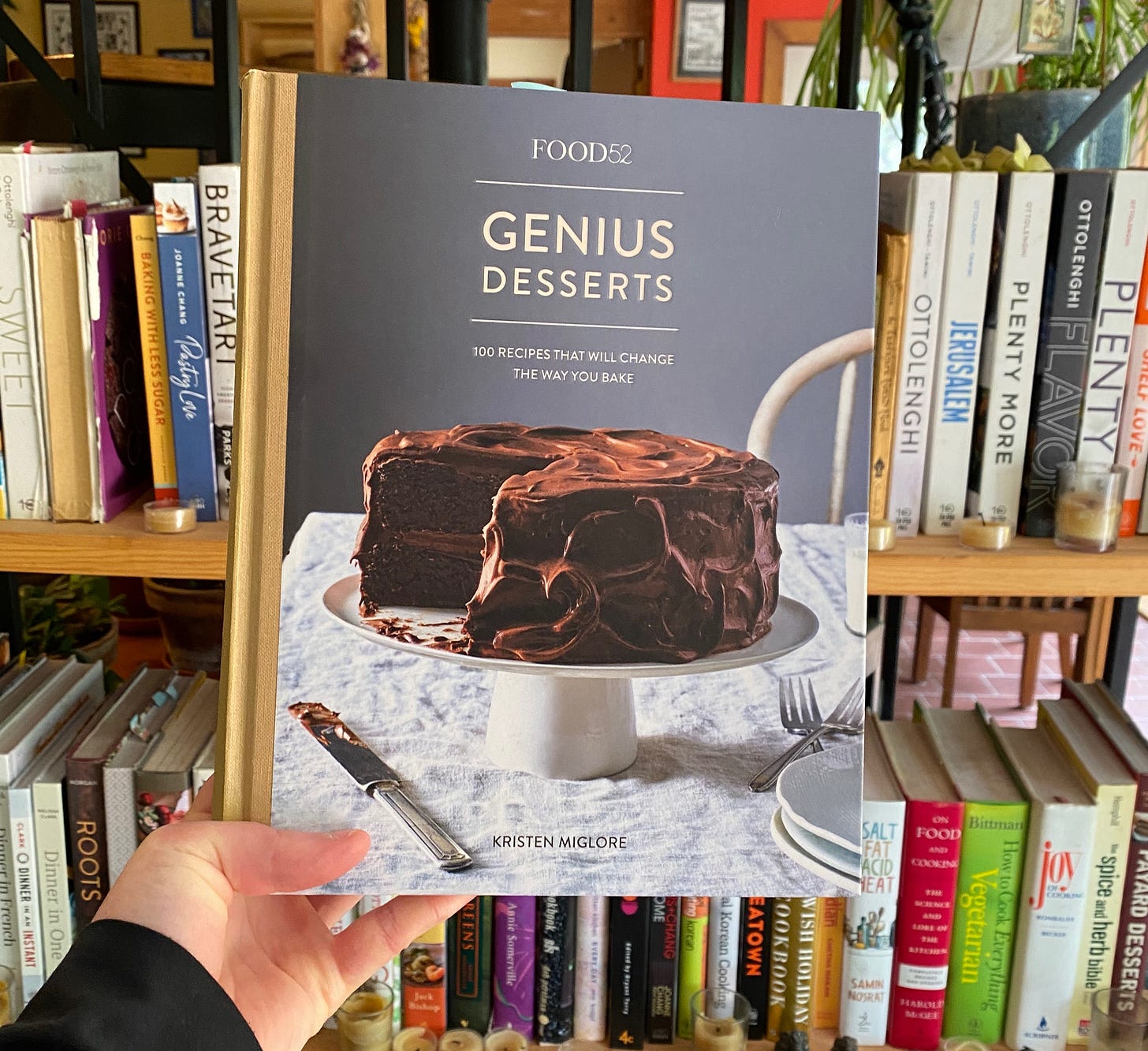 My hand holding up Genius Desserts in front of a shelf of cookbooks.