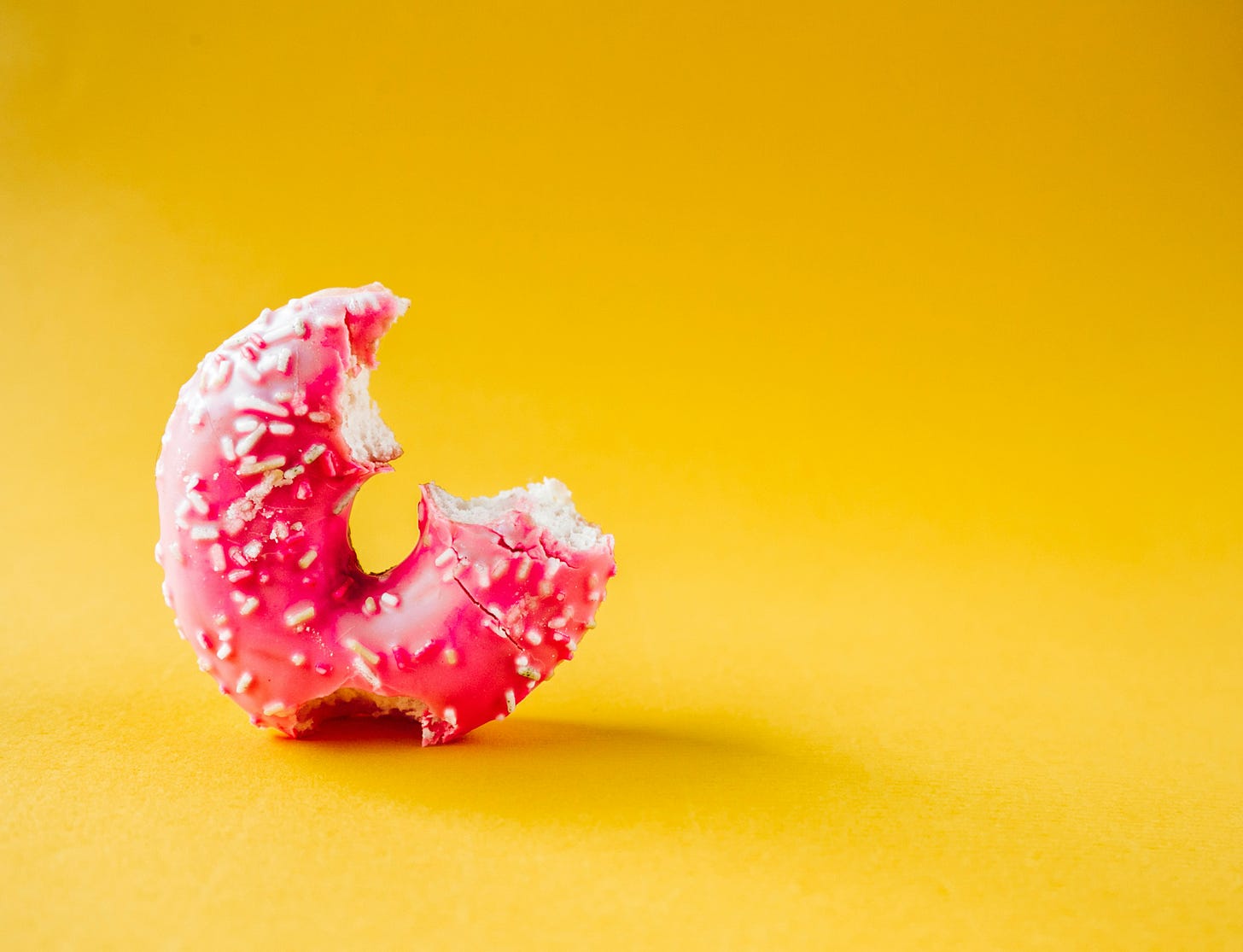 Image of a half-eaten donut with a pink glaze and white sprinkles, against a yellow background