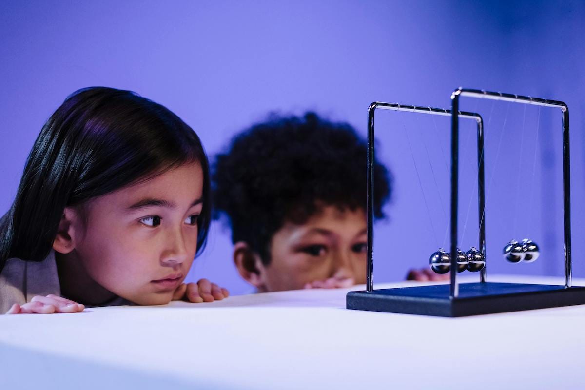 two children stating at a Newton's cradle or pendulum – a device that typical sits on desktops, consisting of five metal hanging balls. when one ball is pulled sideways then released, it collides with the other balls, which forces the ball on the other side into motion, thus starting a back-and-forth chain reaction that, while not literally "perpetual," tends to continue for a very long time.