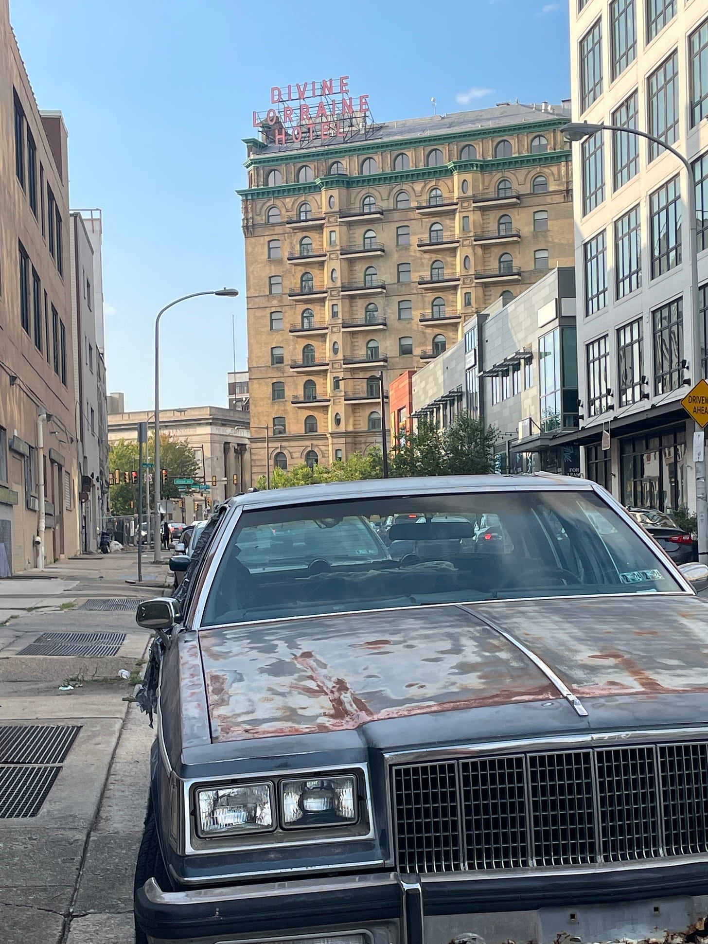 a street scene in Philadelphia, the foreground is a very old rusted sedan parked on the street, in the background rises a victorian high rise with a large sign on the roof in red letters reading "Divine Lorraine Hotel"