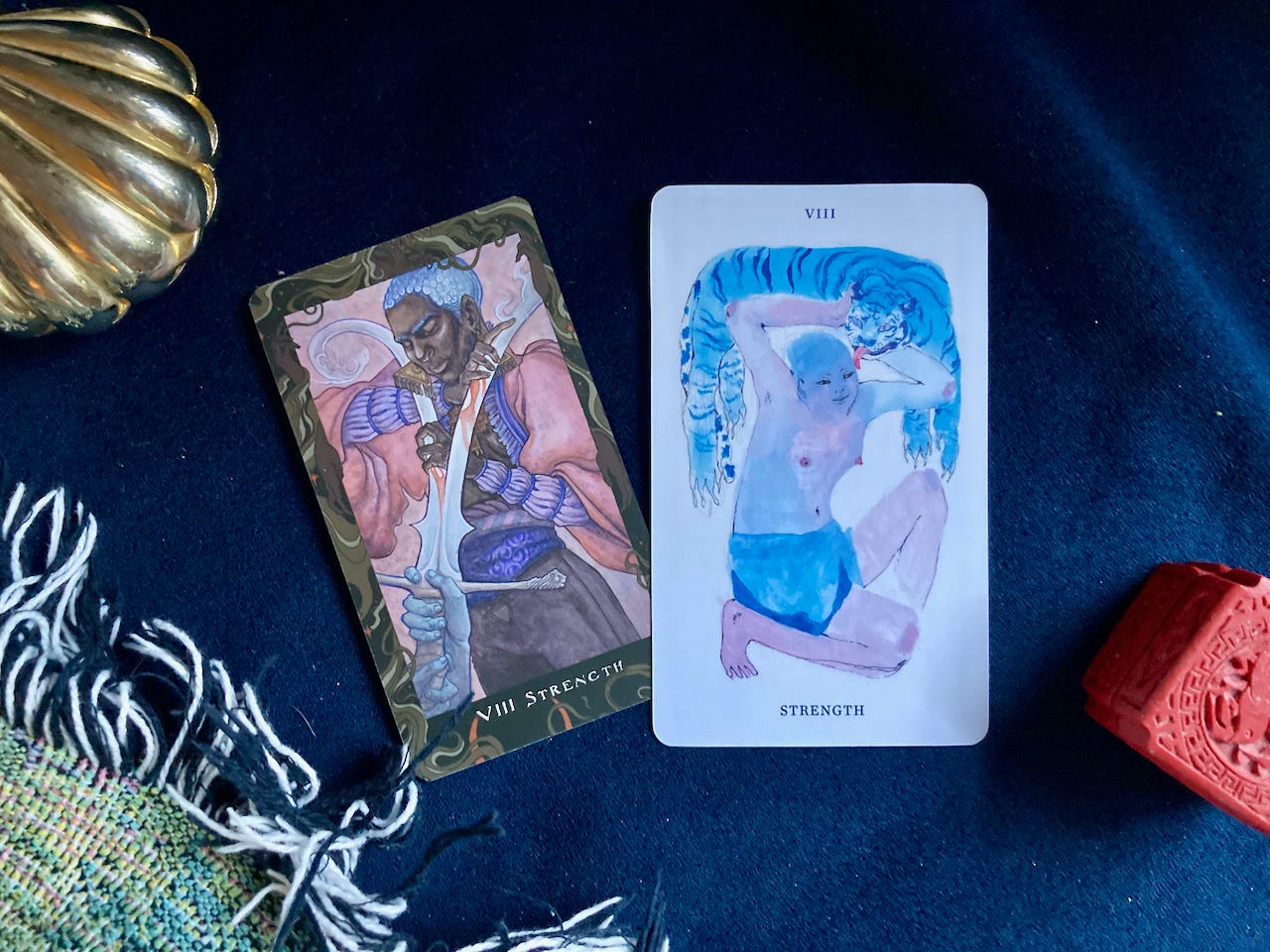 Two Strength tarot cards lay on a dark blue velvet background. One card shows a dark-skinned person with white hair splitting a sword down the middle with their bare hands. The other shows a light-skinned, round-bellied, smiling person kneeling and holding a blue tiger over their head, which licks them.