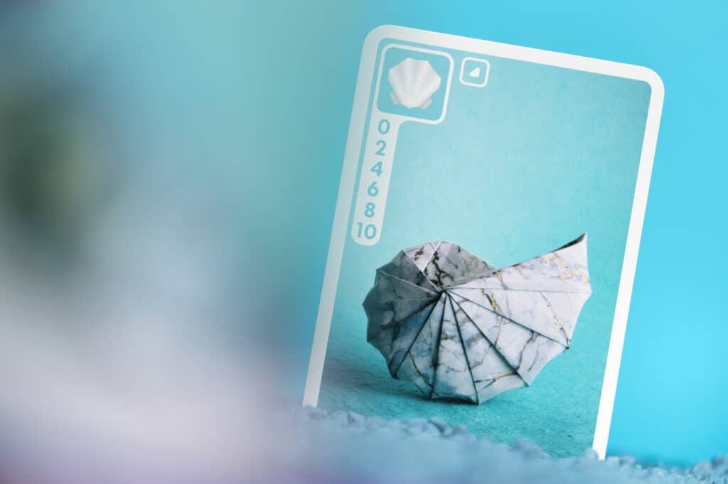 Une carte à jouer, on voit un coquillage en origami sur un fond bleu clair, avec un pictogramme de coquillage en haut à gauche, un symbole permettant d'identifier la couleur de la carte et un barème de points