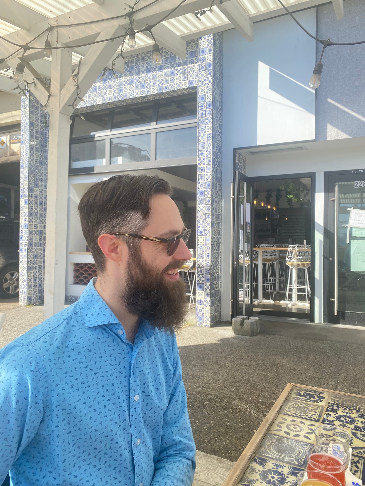 Jeff, at an outdoor table at Cerveceria Astilleros. He's wearing a blue patterned button-down shirt and smiling at someone out of frame across the table. The indoor area of the brewery is visible in the background, blue and white tiles surrounding the open garage door.