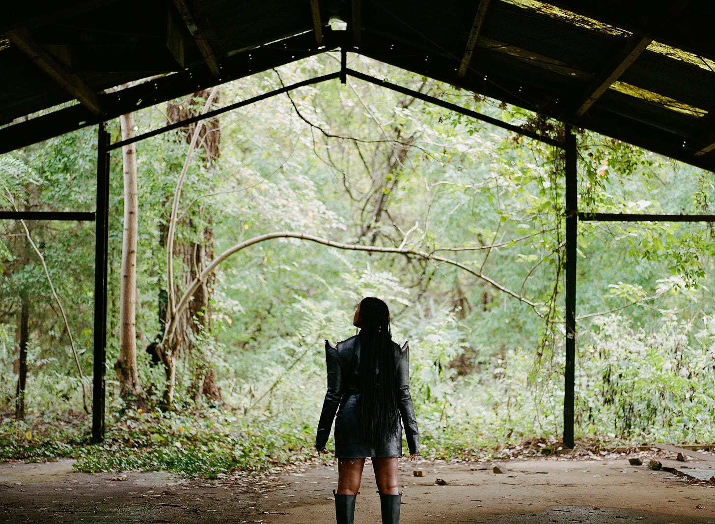 I’m wearing a Zaire Studio mini dress I made with drop cloth canvas painted with black Flex Seal and tall rubber Ganni boots. Buss down middle part braids are almost touching my exposed thighs and I am underneath a decompsing steel structure gazing at the promise of the forest. 