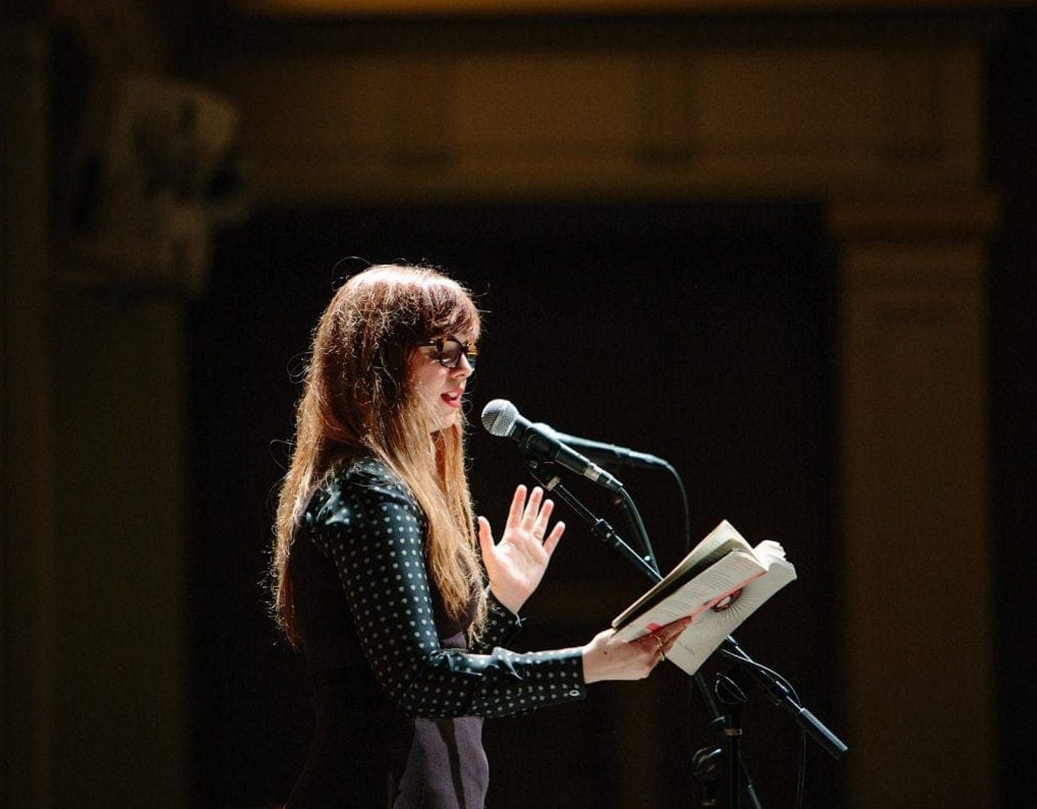 Amber reading from "Dark Sparkler." She speaks into a microphone and holds the book open in front of her. 