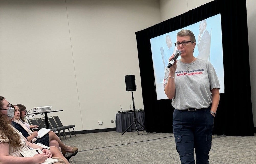 Woman talking into handheld microphone while walking in front of audience; she is wearing a t-shirt that reads "More conversation. Less Polarization." 